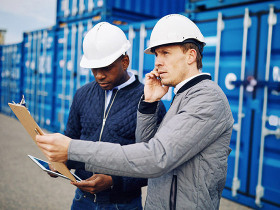 Two men wearing hard hats reviewing documentation with containers in the background