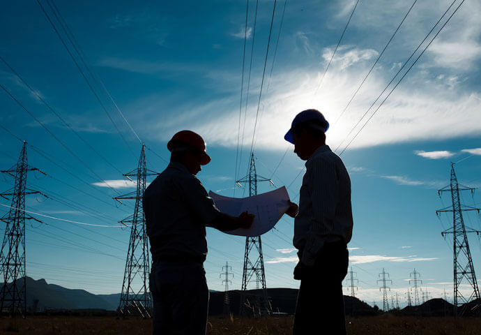 Two men in hardhats reviewing document with power lines in the background